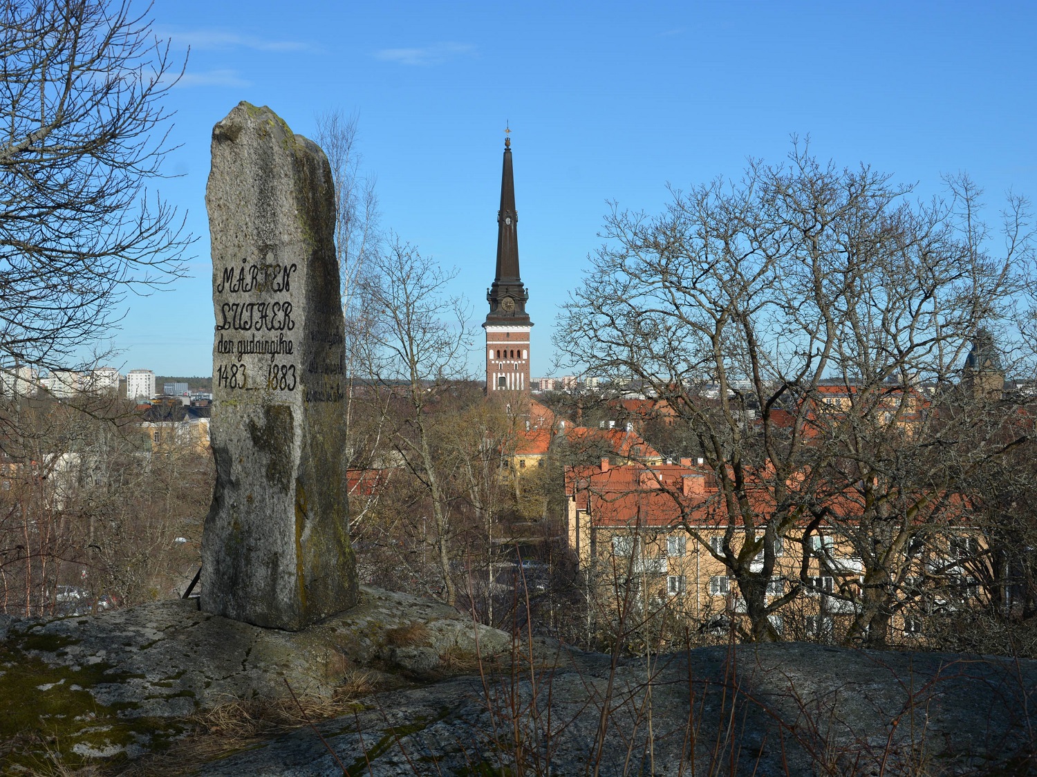Bild med större rest sten med ingravering. Utsikt med blå himmel över Västerås hustak en höstdag.