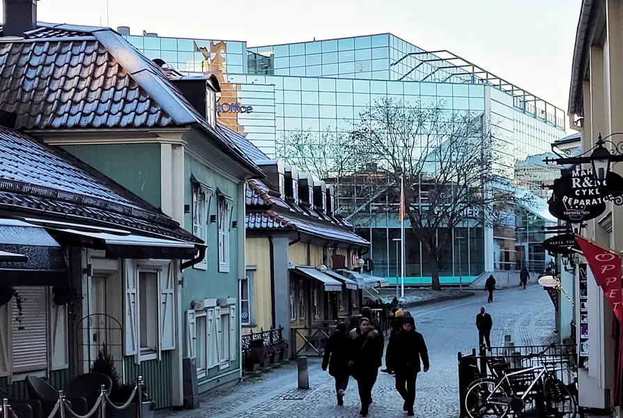 En
kullerstensstadsgata med olikfärgade trähus, människor och cyklar, i bakgrunden
syns ett lite större glashus.