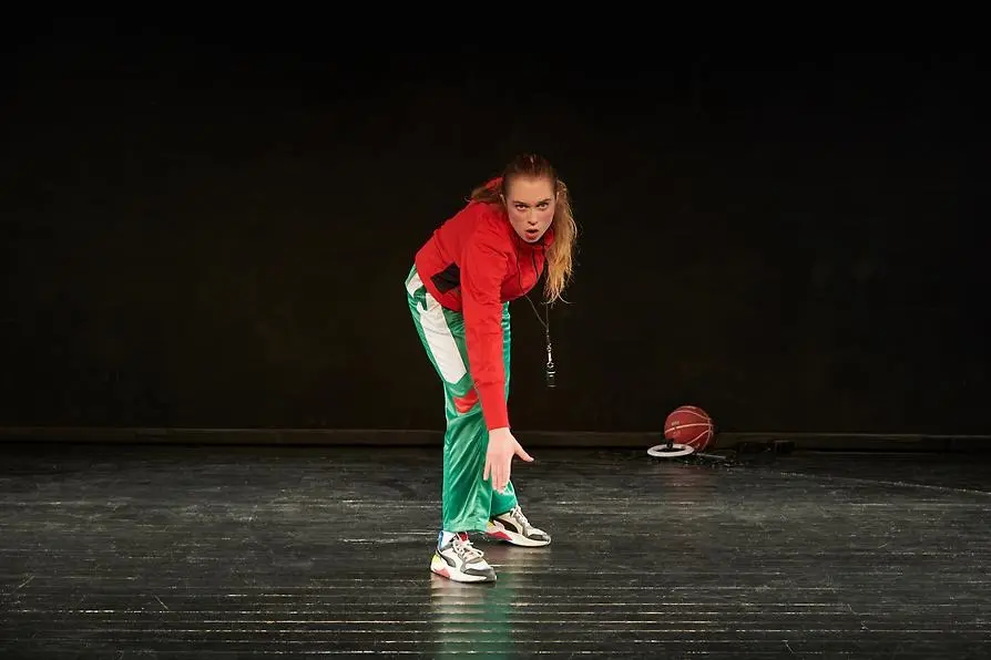 En kvinna med långt halvblont hår, joggingskor, gröna träningsbyxor och röd träningsjacka står halvt framåtlutad, redo att springa. Golvet och bakgrunden är svarta, en bit bakom henne ligger en basketboll.