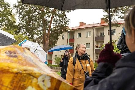 Karin Nordström står inför en grupp och pratar. De står framför ett putsat hus från 40-60-talet. Det regnar och många har uppfällda paraplyer.