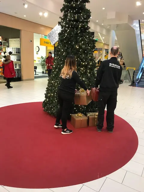 Två personer lägger julklappar vid en stor julgran i shoppingcentret.