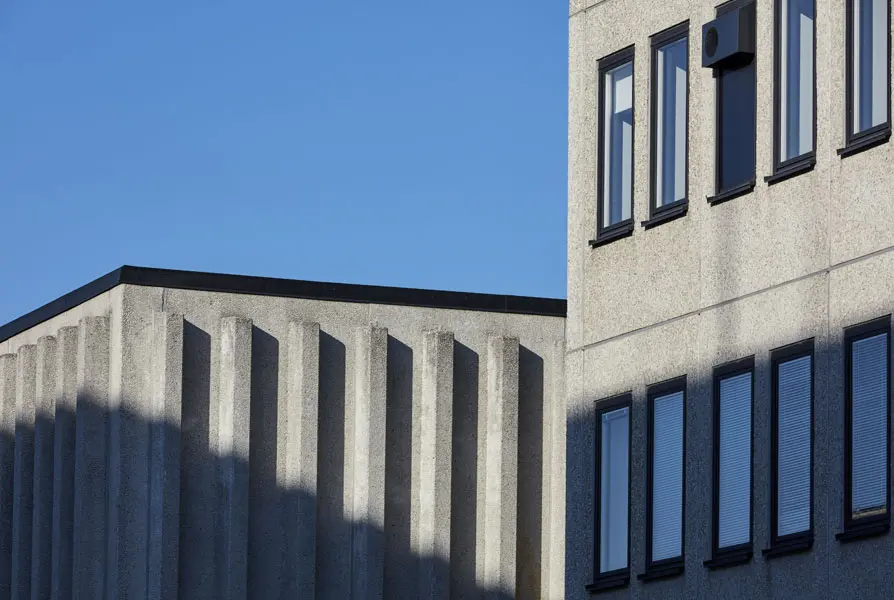 Närbild på de två olika fasaderna med utskjutande betongprofiler i den lägre byggnaden och släta betongelement med rader av fönster i den andra. Foto: Lasse Fredriksson