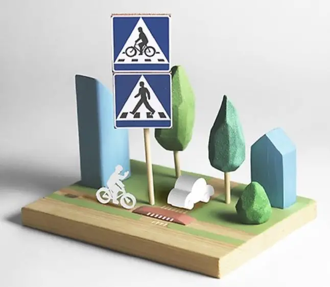 Bild på leksaksklossar av en väg, några hus och träd, en stolpe med skyltar för övergångsställe och cykelöverfart. En vit pappfigur cyklar över vägen och en vit bil står och väntar på vägen.