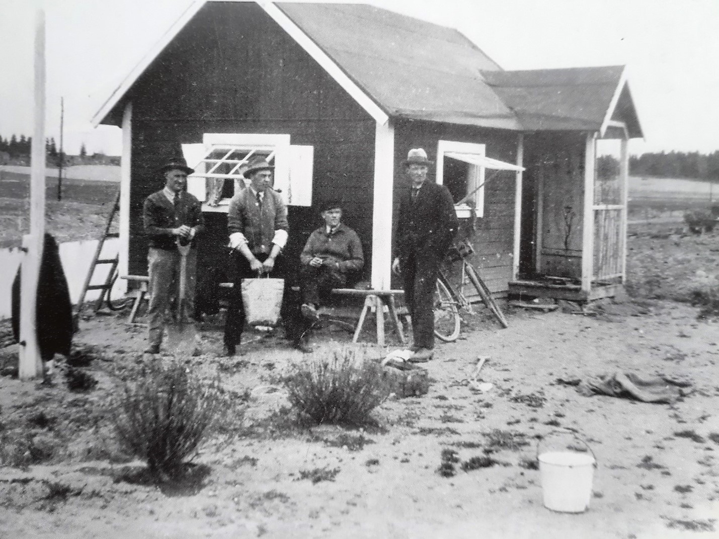 Svartvit bild på kolonistuga med 4 män i hatt poserande utanför.