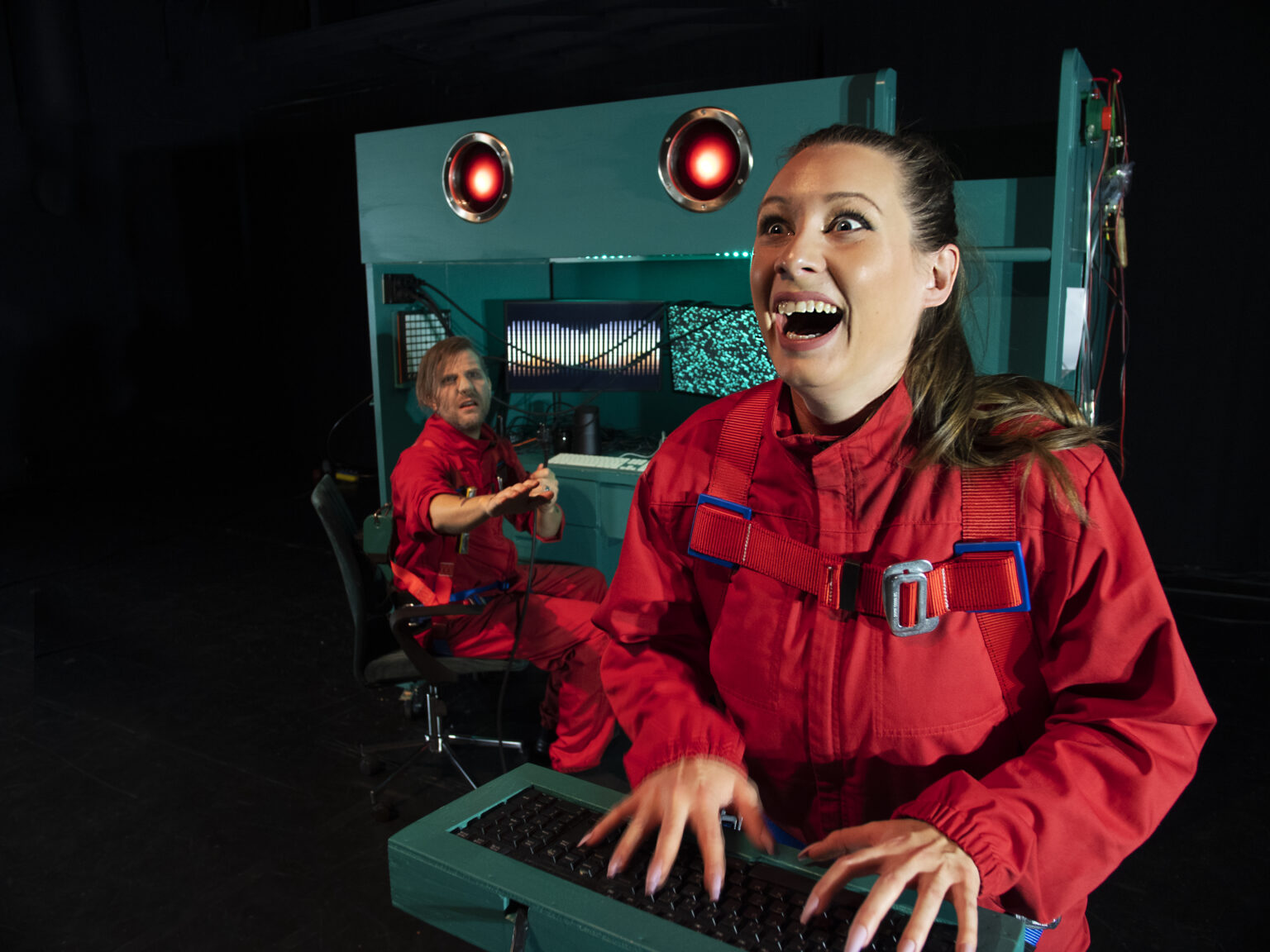 I förgrunden syns en person med röd arbetsoverall, som ser glad ut och sitter med båda händerna på ett tangentbord. I bakgrunden sitter en annan person med röda arbetskläder, vid en grönblå maskin med två lysande lampor som ser ut som ögon.