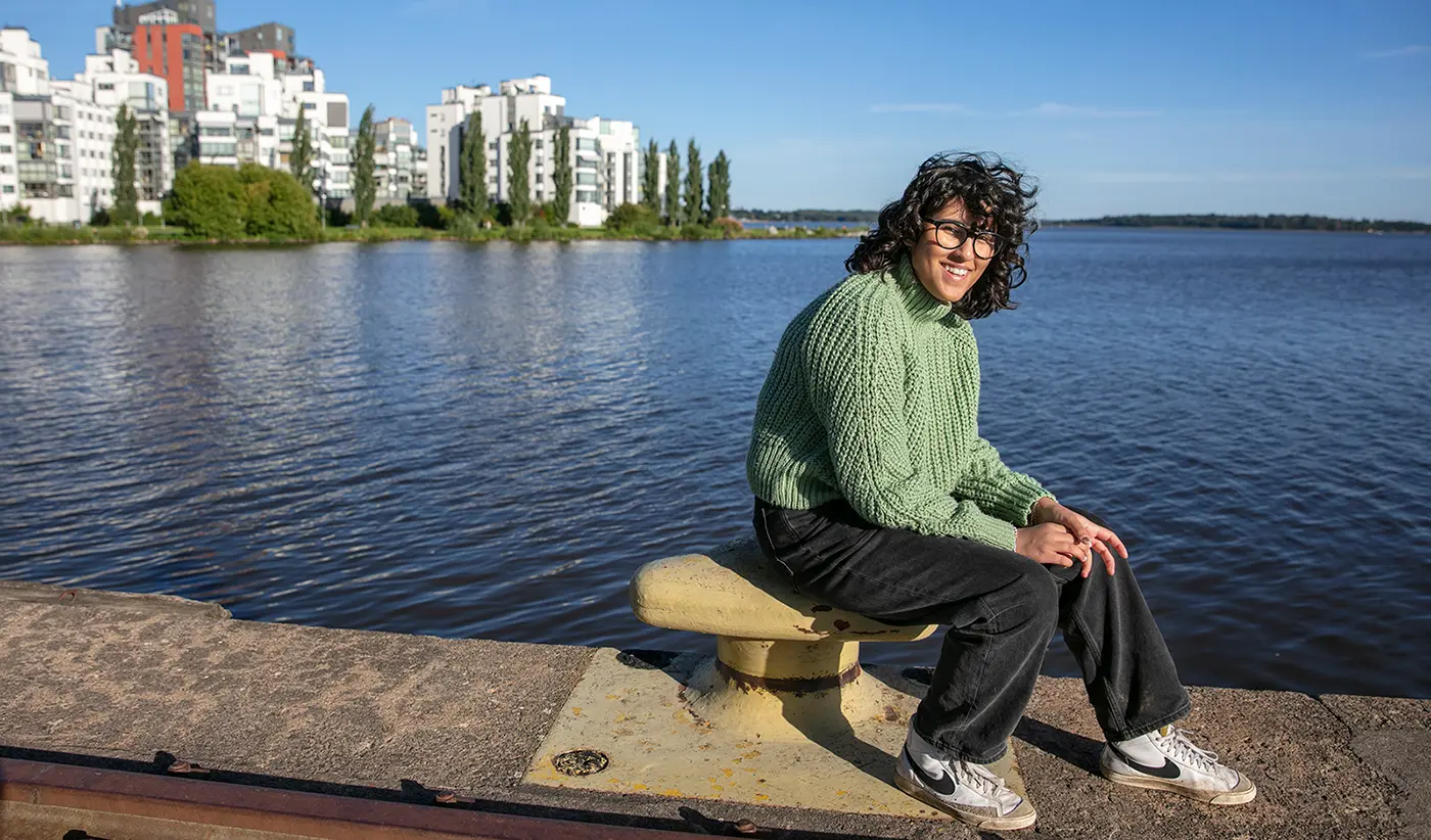 En ung kvinna sitter på en pir framför Lillåudden i Västerås. Man skymtar vatten och byggnader i bakgrunden.