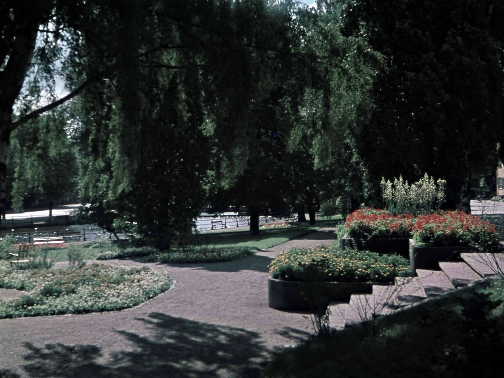 Park med lummiga träd, stor grusgång och en trapp med röda blommor i en plantering intill.