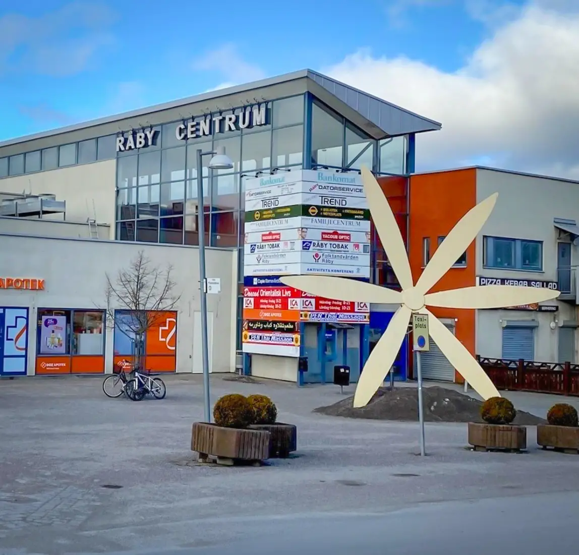 Gallerian i Råby centrum har en ljusgrå putsad fasad längst ner och höga glaspartier högst upp. På ena sidan står texten Råby centrum. I förgrunden syns en stor dekoration i form av en ljusgul blomma med sex kronblad. Det står två cyklar lutade mot ett träd.