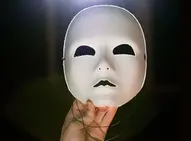 Hand som håller upp vit mask föreställande ett ansikte. 