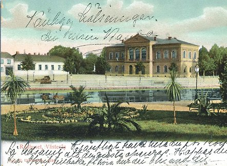 Vykort i färg med en handskriven hälsning. Motivet är Rådhuset som det såg ut cirka år 1900. Vyn är tagen från Stadsparken, med en park med palmer samt Svartån i förgrunden.